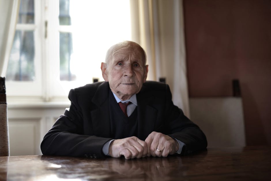 A serious elderly man in formal wear sitting at a table indoors, captured during the day.