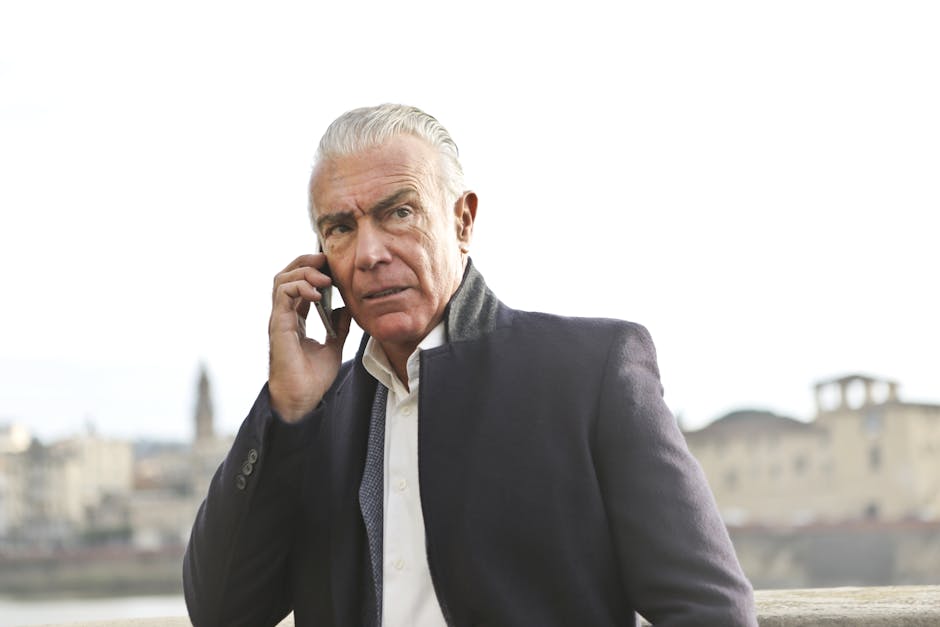 Senior man with grey hair in formal attire using a smartphone outdoors against a city backdrop.