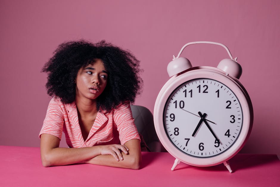 Young woman leaning on table with pink alarm clock, looking pensive.