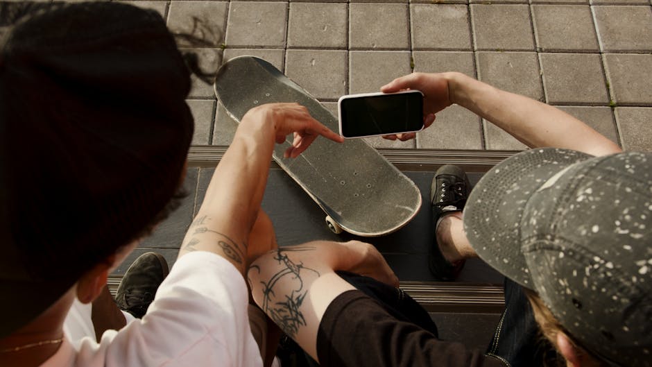 Two teenagers sitting outdoors with a skateboard, looking at a smartphone, enjoying leisure time together.
