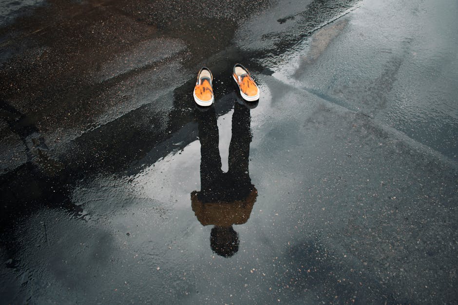 Reflection of legs and shoes in a puddle on a wet street, creating a surreal urban scene.