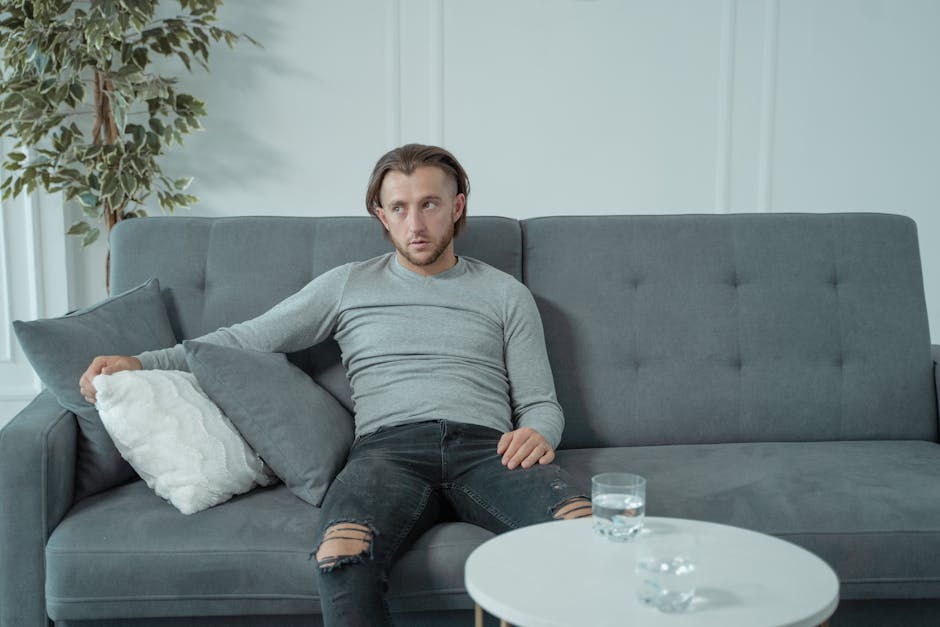 A man in casual attire sits on a gray sofa, appearing thoughtful and relaxed in a modern living room.