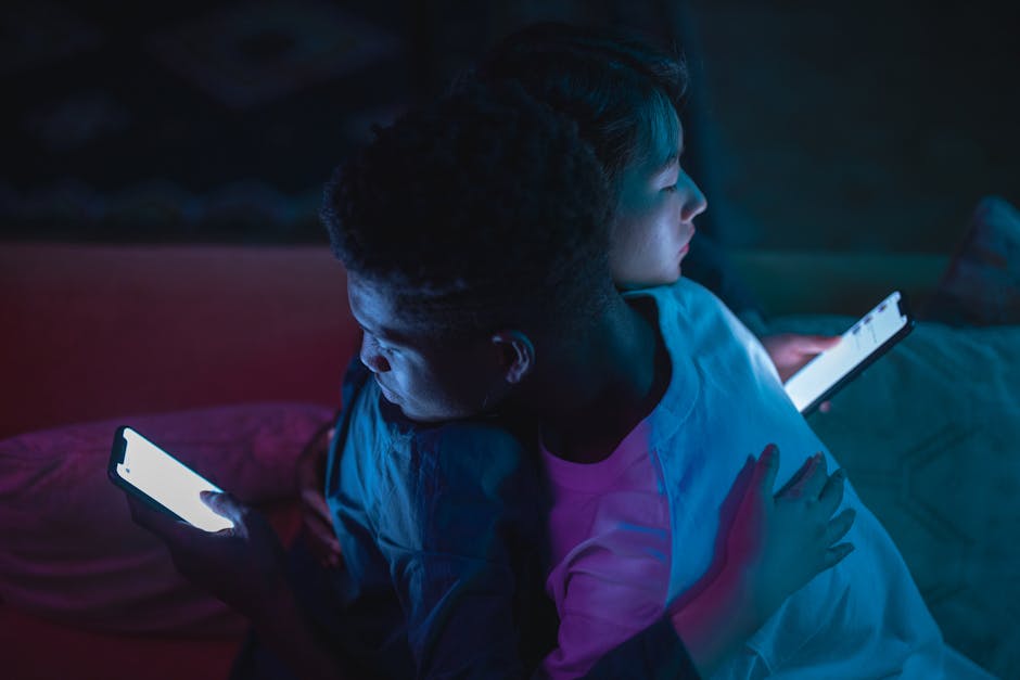Interracial couple embracing while using smartphones, casting a warm glow in a dark room.