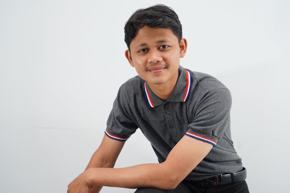 A confident young man in a casual shirt smiles warmly in a bright studio setting.