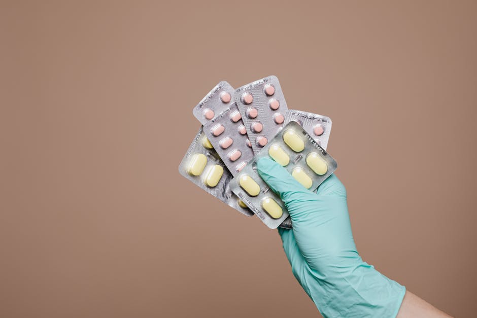 Close-up of a gloved hand holding multiple blister packs of tablets against a neutral background.