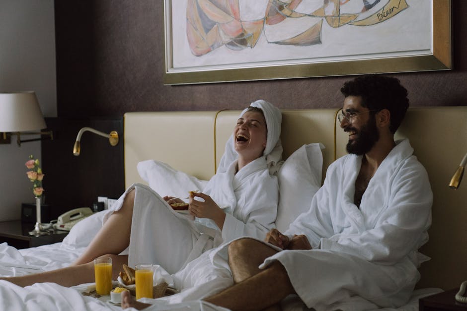 A joyful couple in bathrobes laughing while having breakfast in bed indoors.
